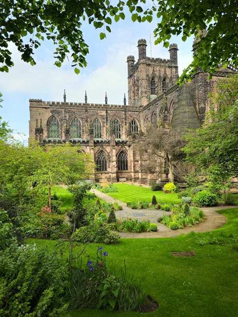 A lush, well-maintained garden in front of a historic stone cathedral showcases neatly trimmed grass, vibrant flower beds, and a variety of shrubs and small trees. The garden features curved pathways 