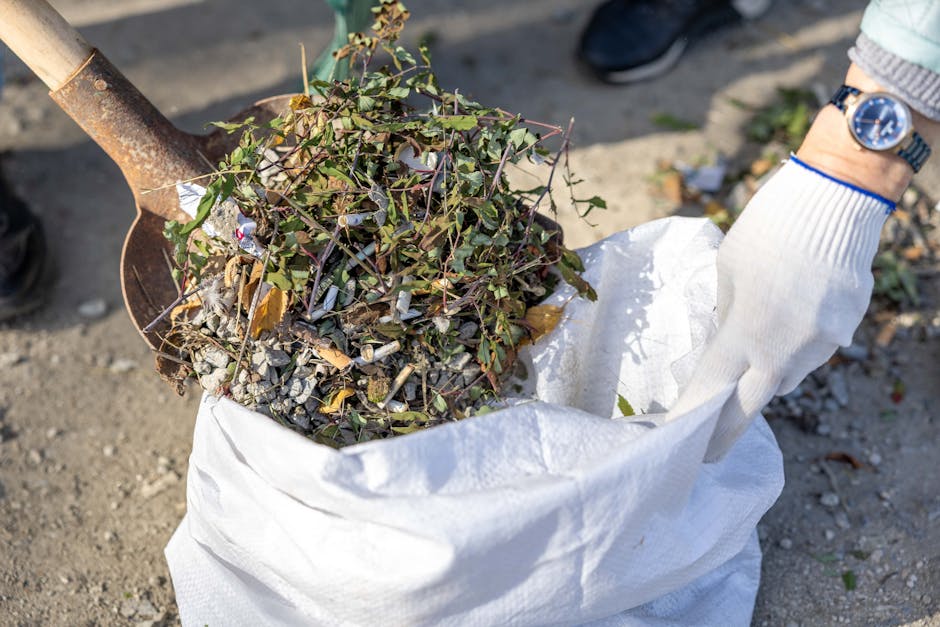 A gardener wearing a white glove is holding a white sack filled with garden waste, including small branches, leaves, and twigs. The scene shows a gravel pathway in a garden setting, with a metal spade